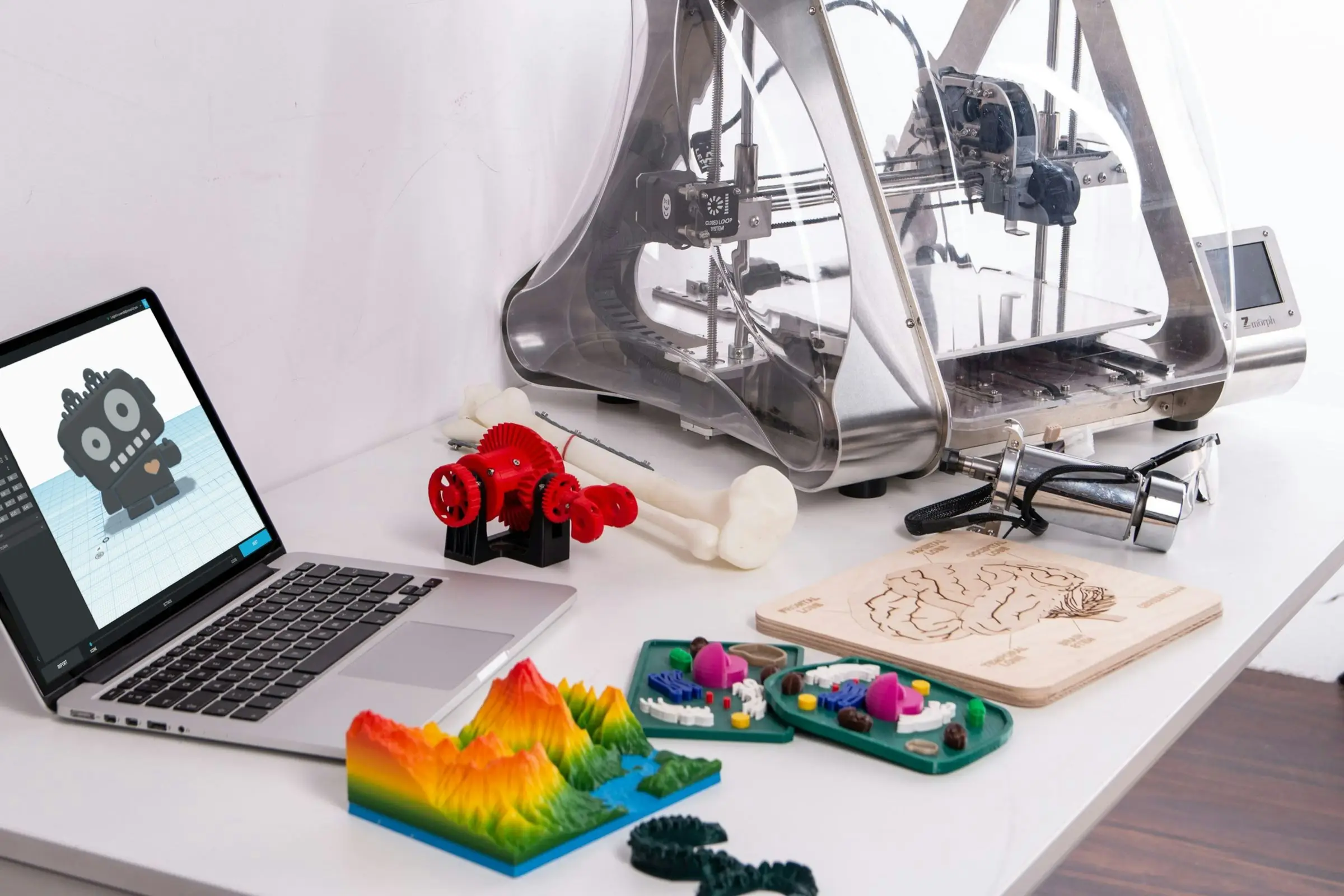 A white desk with a laptop showing a 3D model, an enclosed 3D printer and several colourful printed models including a terrain map and a bone.