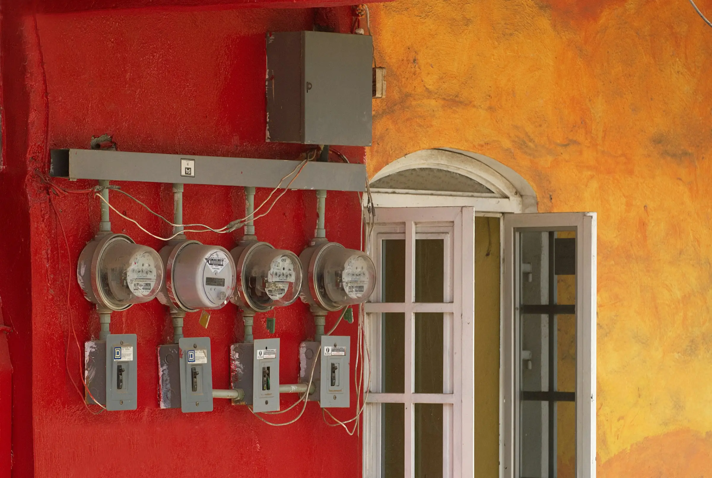 Four electricity meters with breaker boxes mounted on a bright red wall beside a white window.