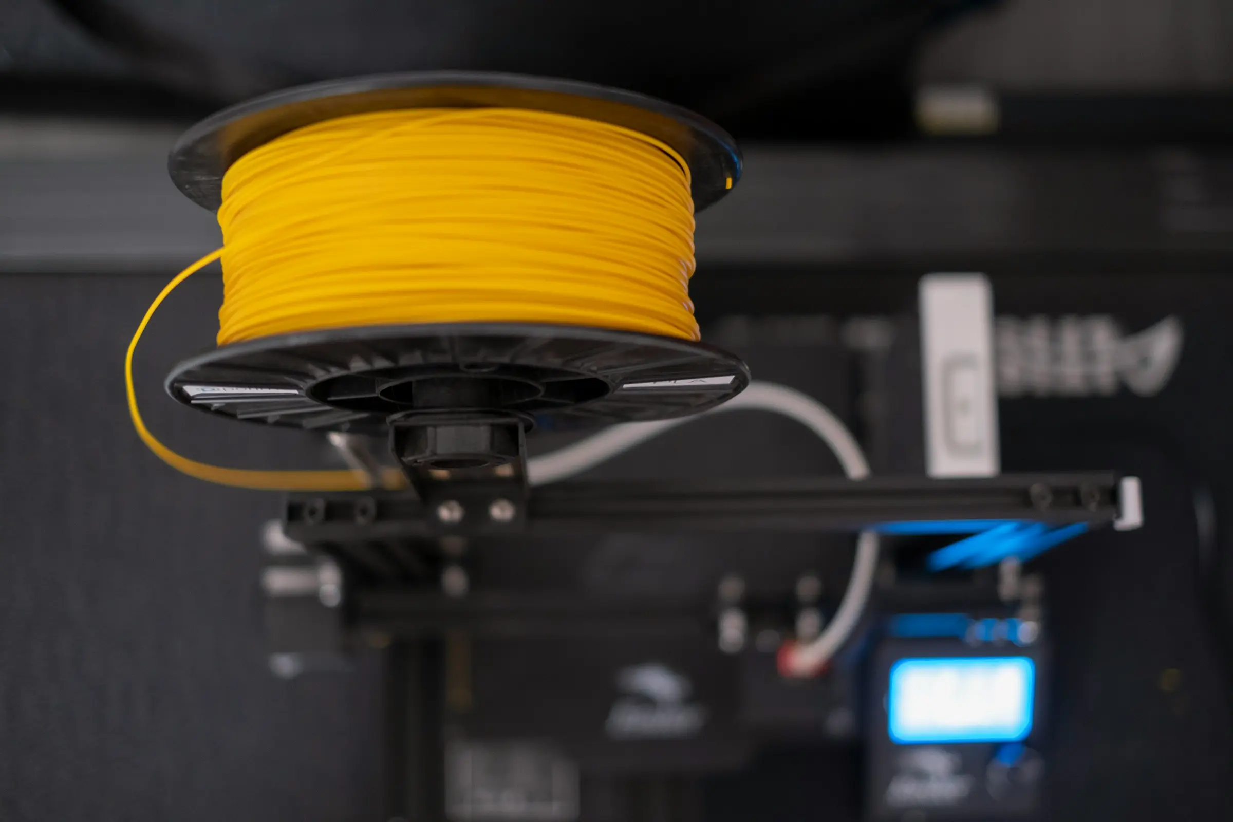 A spool of yellow filament on a holder above a 3D printer, with the printer's blue screen blurred below.