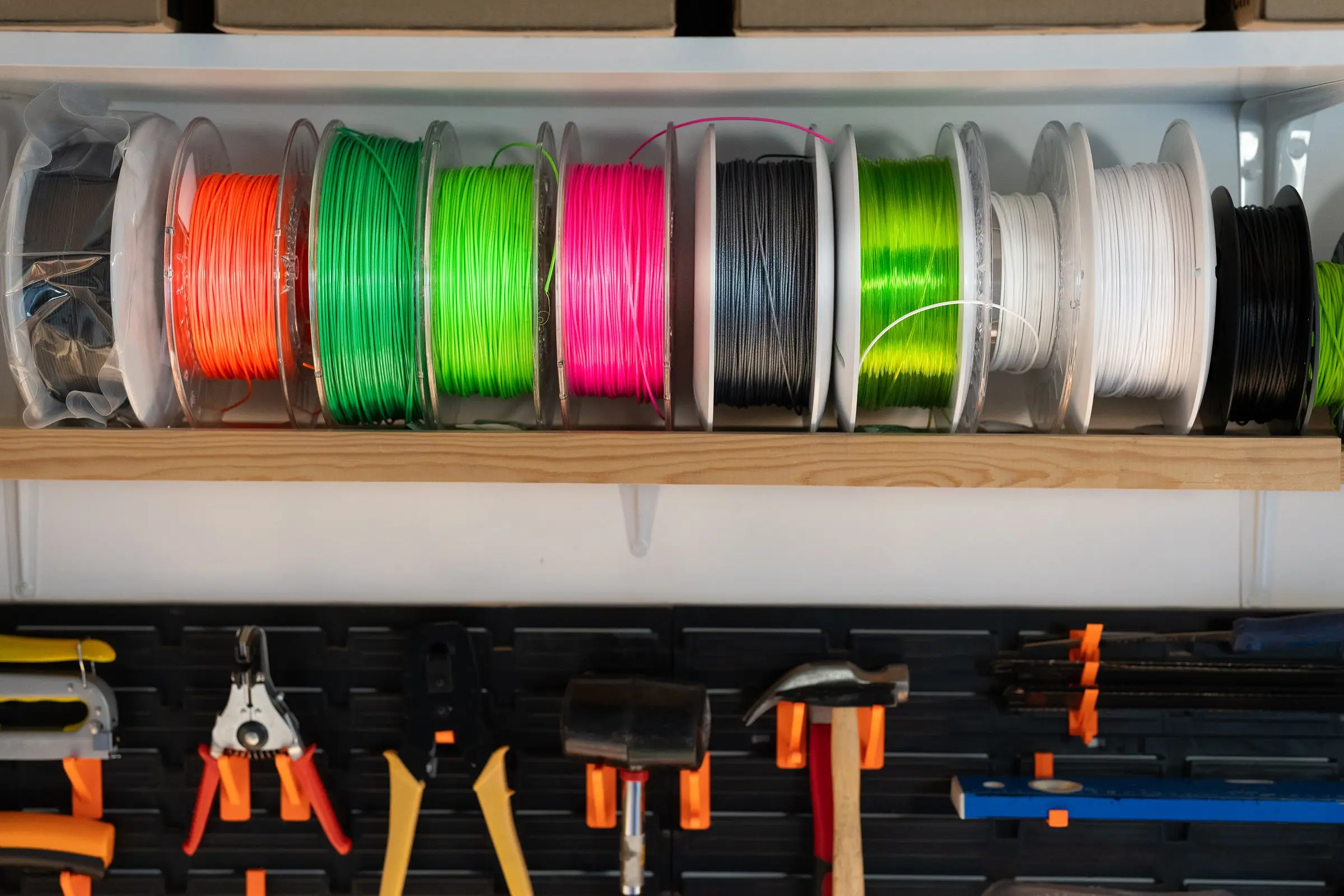 A shelf of filament spools in orange, green, pink, black and white above a tool wall with pliers and hammers.