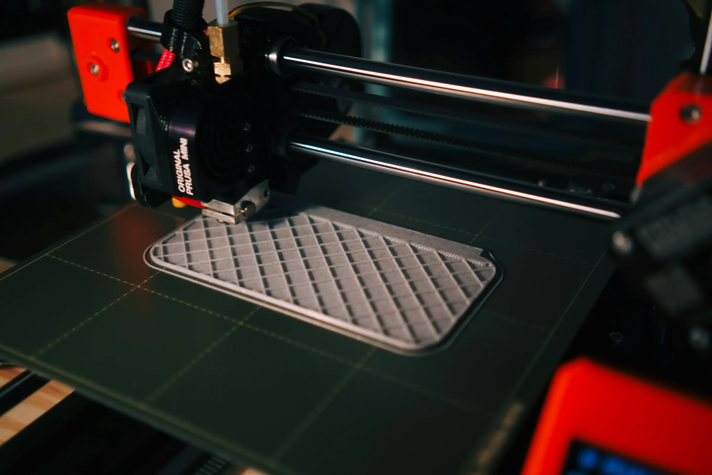 A 3D printer laying down a flat grey panel with a diamond grid pattern on a dark print bed.