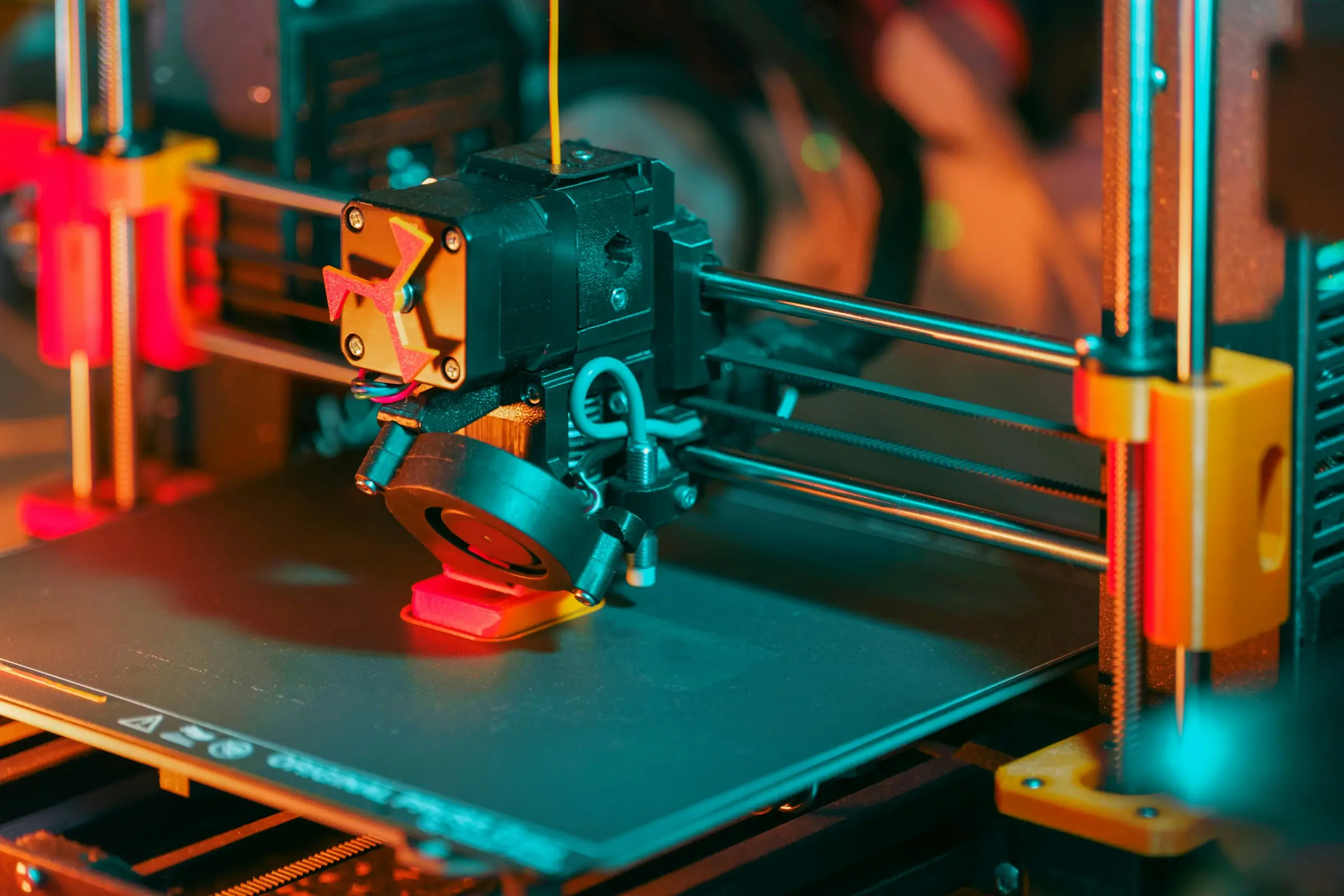 Close-up of a 3D printer extruder and part cooling fan printing a small red object.