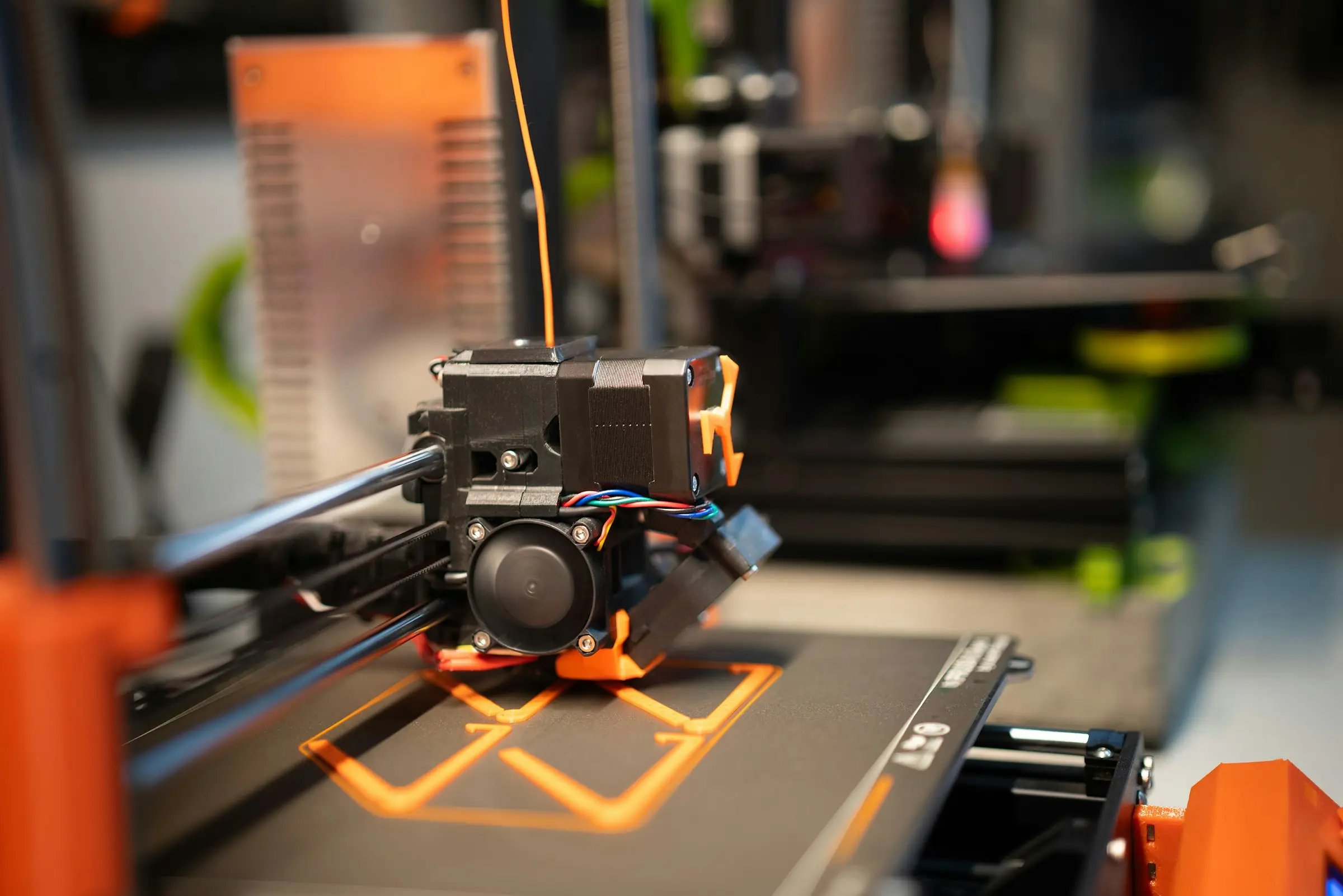 A 3D printer laying the first layer of an orange outline on a dark print bed.