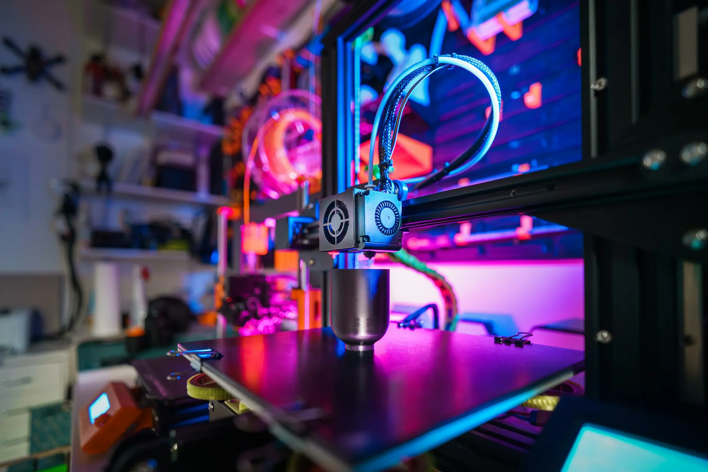 A 3D printer print head with two cooling fans above an empty bed, lit in pink and blue, with workshop shelves behind.
