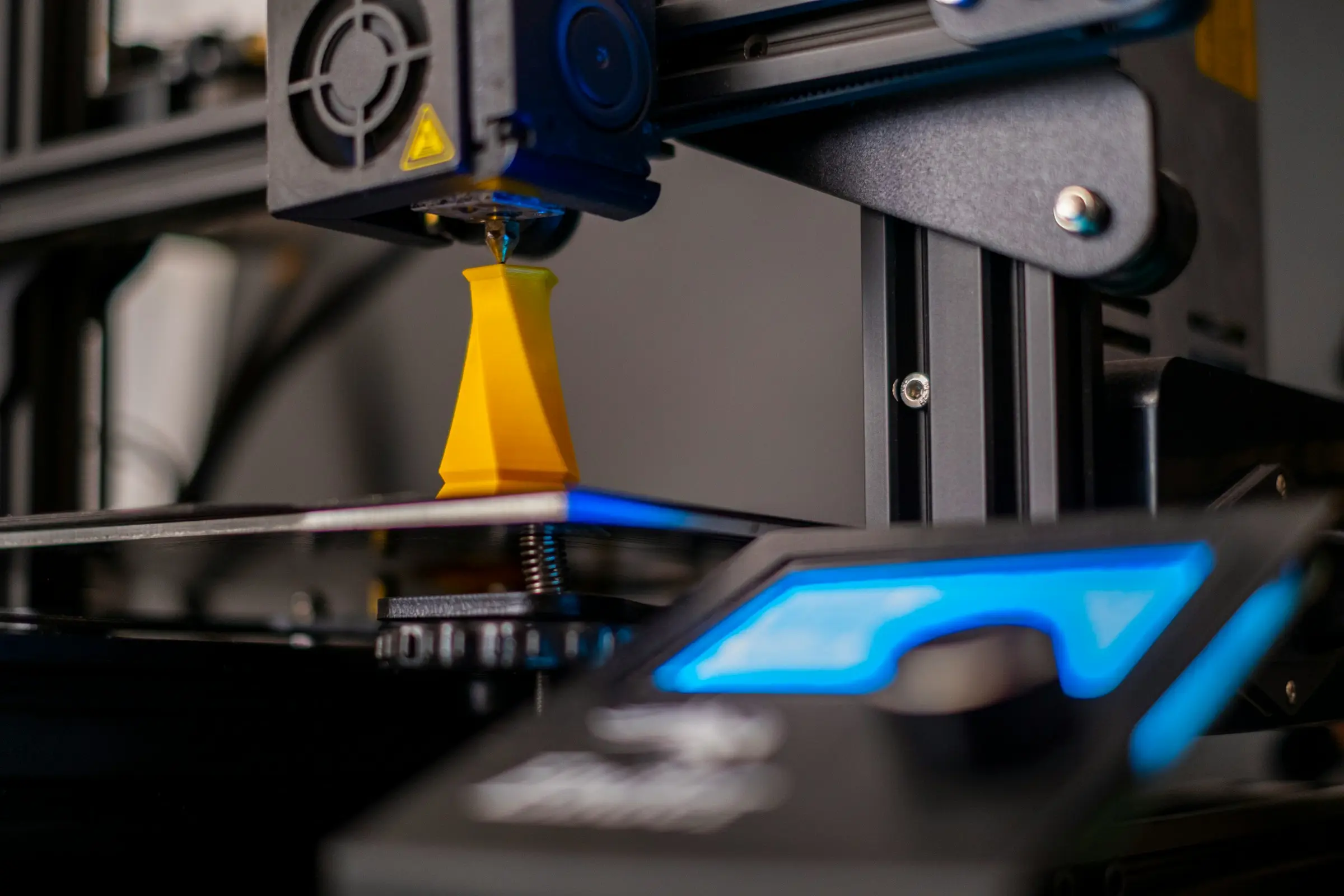 A 3D printer nozzle finishing a tall yellow faceted vase, with a blue-lit control screen blurred in the foreground.
