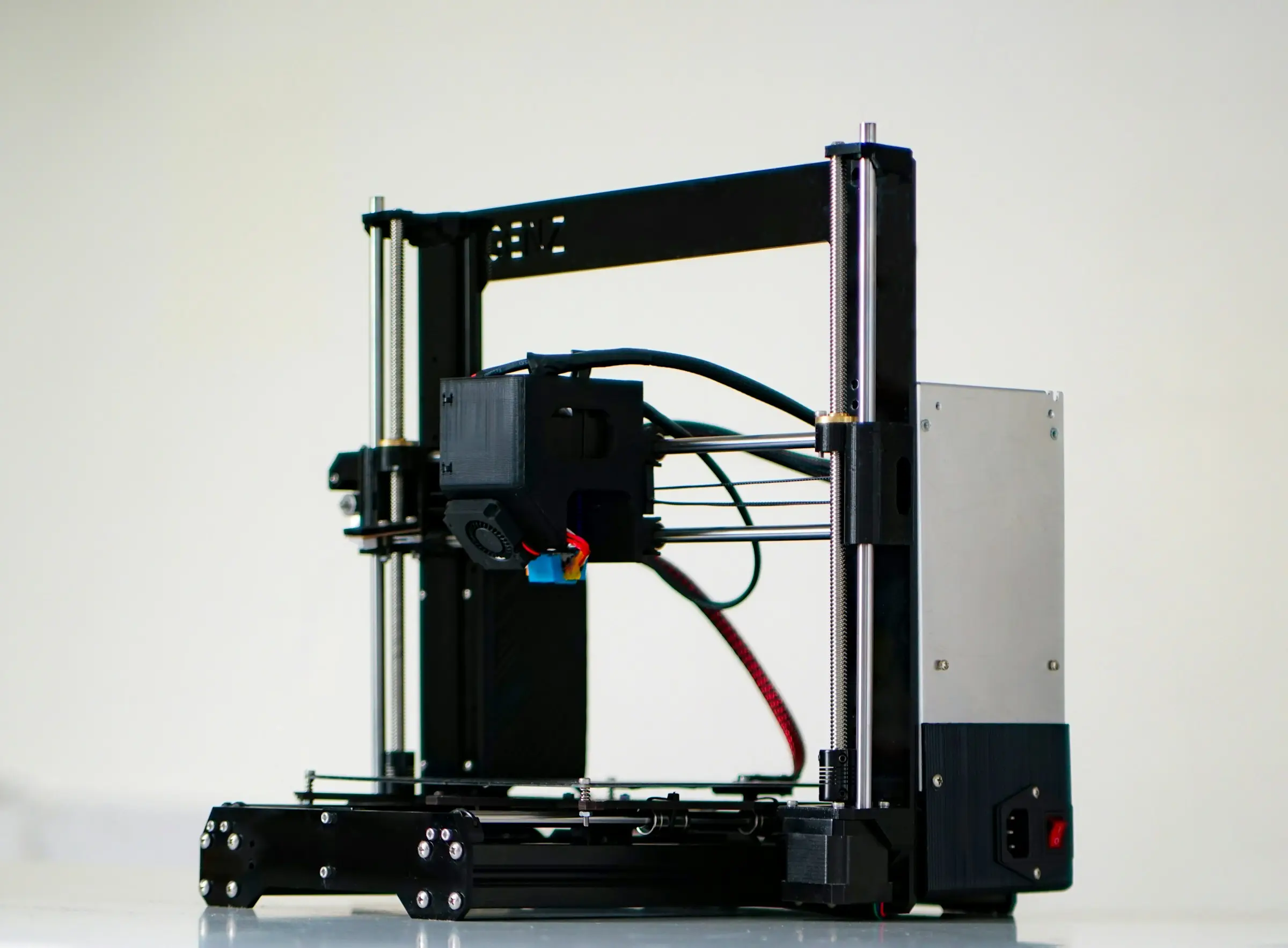 A black open-frame 3D printer with its power supply on the side, photographed against a plain white background.