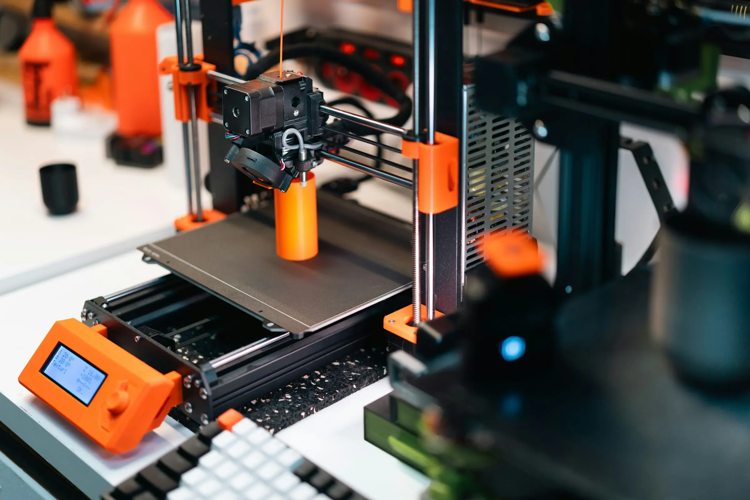 An open-frame desktop 3D printer printing a tall orange cylinder on a desk. A stock photo, not either Bambu Lab model.