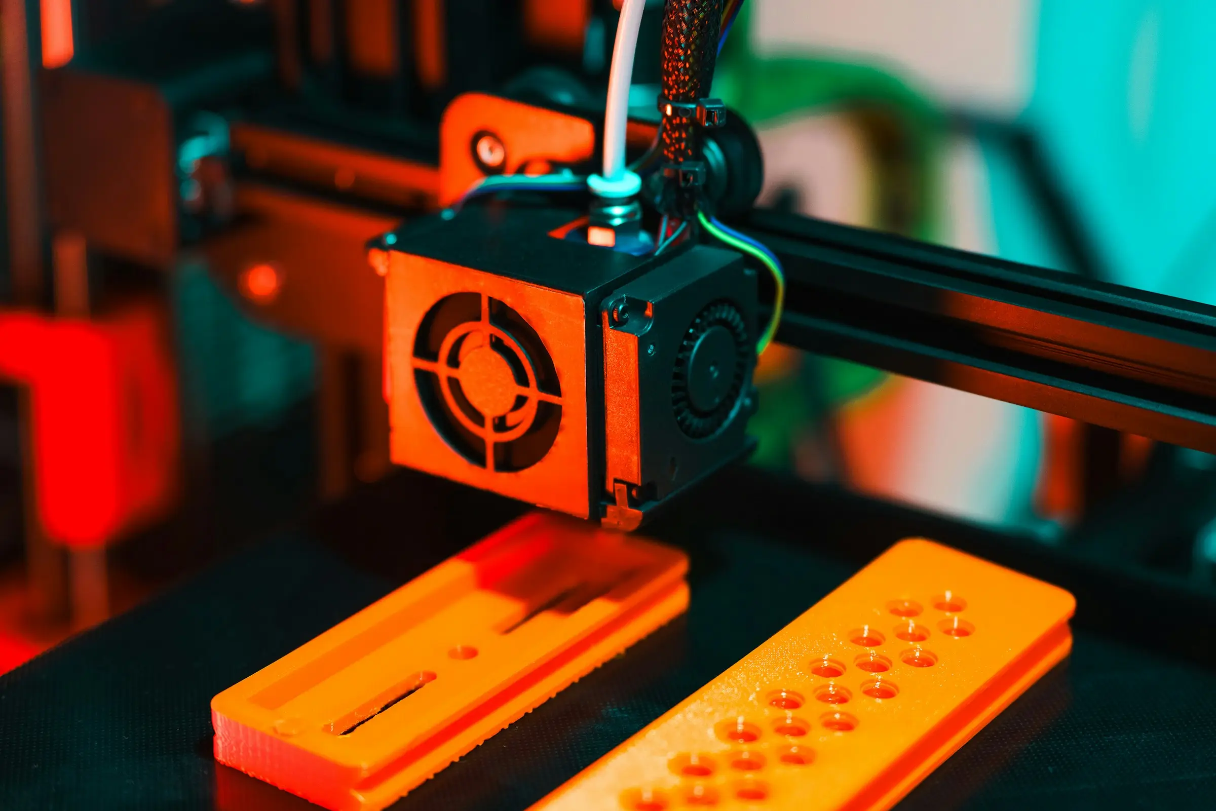 A 3D printer print head above two flat orange parts with slots and holes, lit in orange and teal.