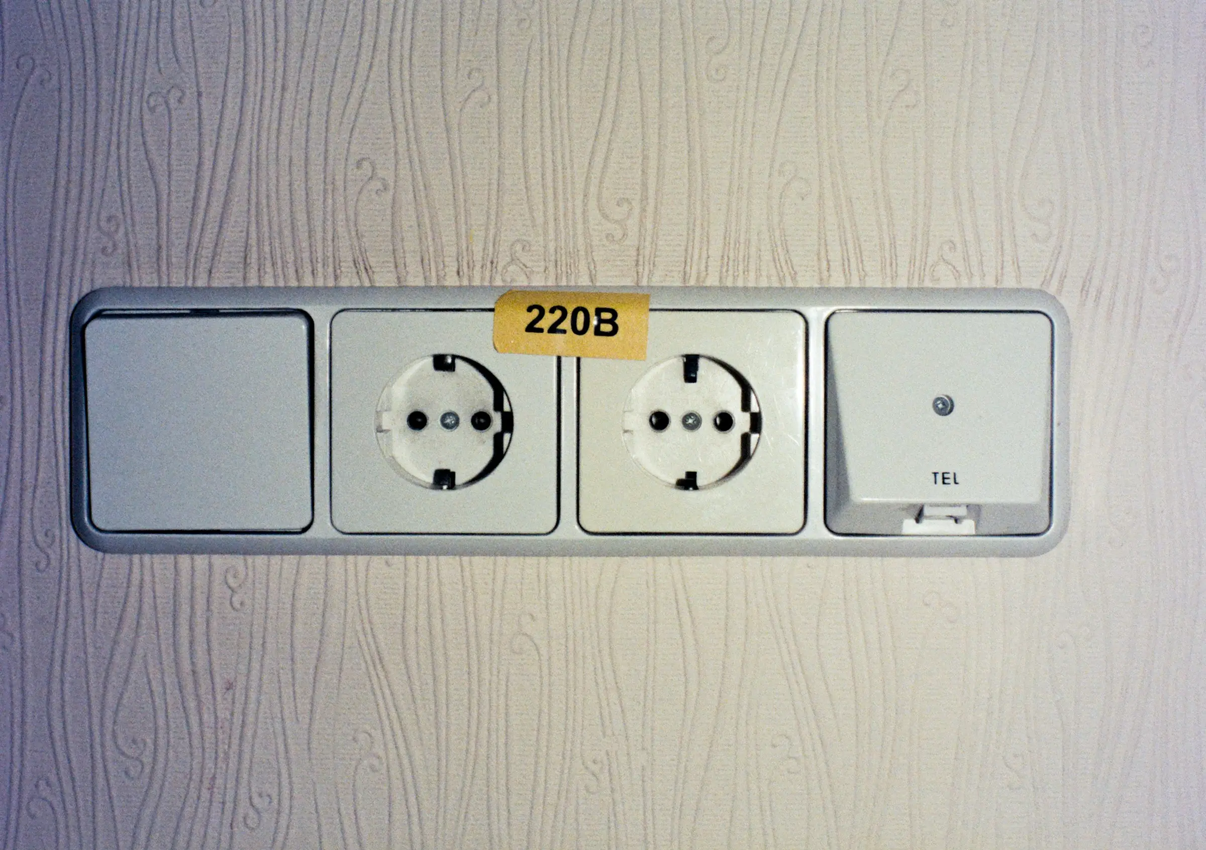 A wall plate with a switch, two round European sockets under a yellow voltage label and a telephone outlet, on textured wallpaper.